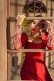 Woman in a red dress with decorative sleeves standing behind a wooden lattice.