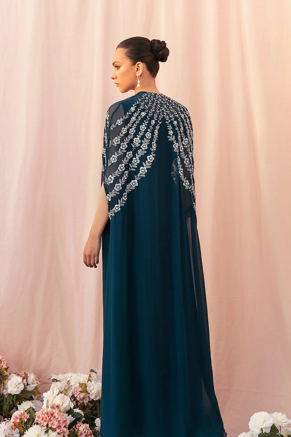 Woman wearing a teal dress with floral embellishments against a pink curtain background.