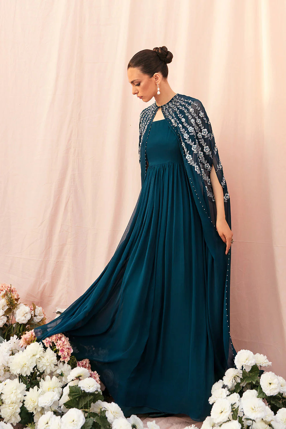 Woman in a teal evening gown with floral embellishments standing among flowers against a pink curtain.